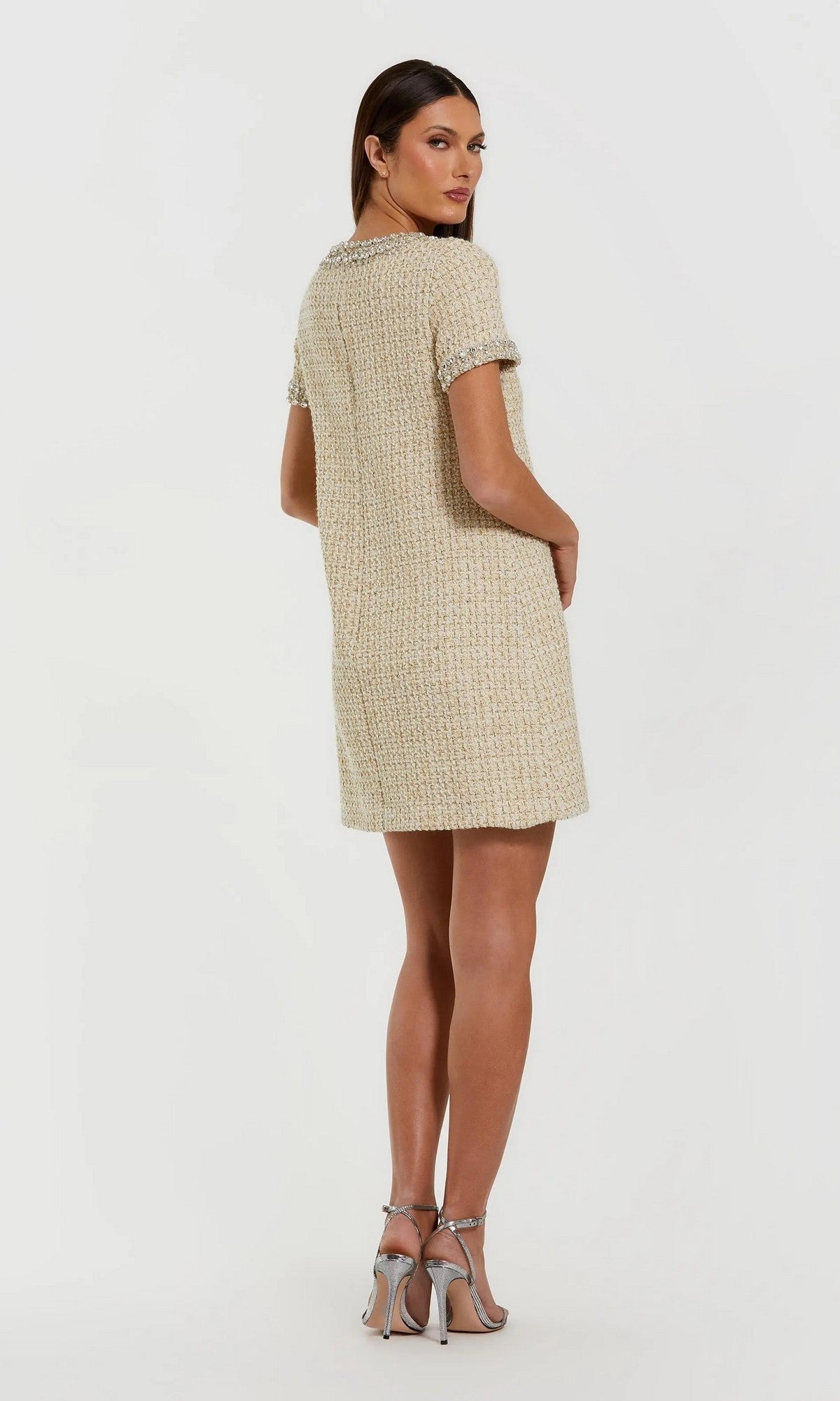 A woman poses against a plain light background, her body turned slightly away, wearing the Mac Duggal 37075 Short Semi-Formal Dress. The boucle mini dress features subtly textured beige fabric, delicate rhinestone beading on the short sleeves for an effortlessly chic appeal, and is styled with silver high heels.